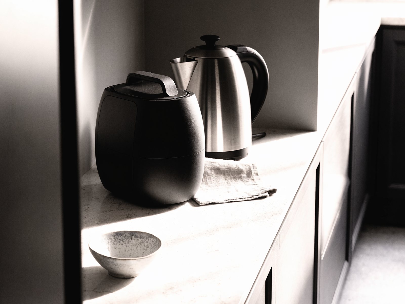 A matte black bean-to-cup coffee machine and a stainless steel kettle on a sunlit marble kitchen worktop